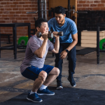 man helping older man perform squats holding dumbbells